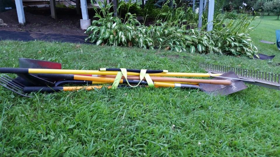 A bundle of shovels, rakes and a fork cinched together by a hi-vis green Shovel Sling, lying on a lawn